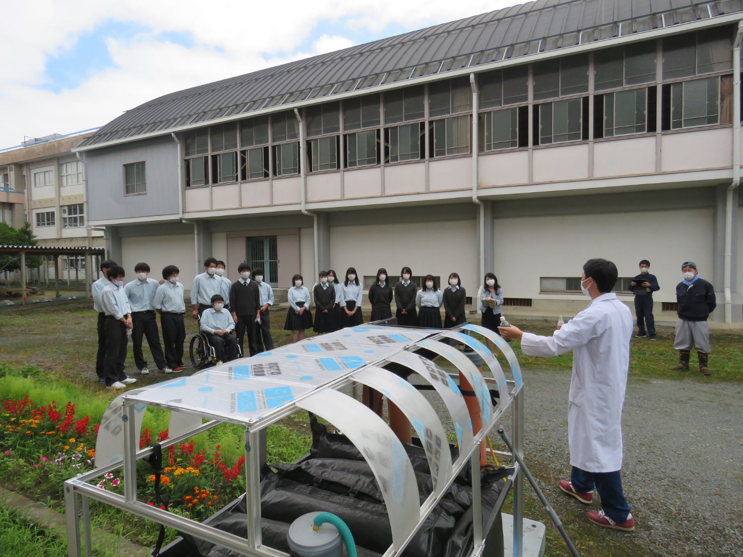 屋外で白衣を着た野添さんがホームバイオガス装置の前で学生たちに説明をしている様子の写真