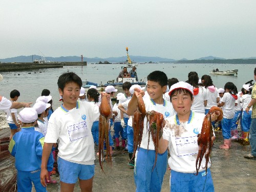 体操服を着た多くの子どもたちが、港の岸壁で水揚げされたと思われる大きなタコを誇らしげに掲げ、笑顔を見せているふるさとの海に親しむ会の写真
