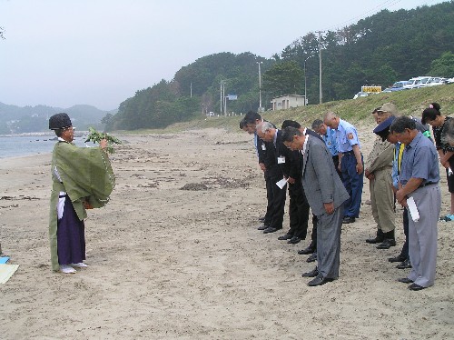 海岸の砂浜で、緑の装束を着た神職が玉串のようなものを持って立っており、それに向かってスーツや制服姿の数人の人々が深々と頭を下げている長須賀海水浴場海開きの様子の写真
