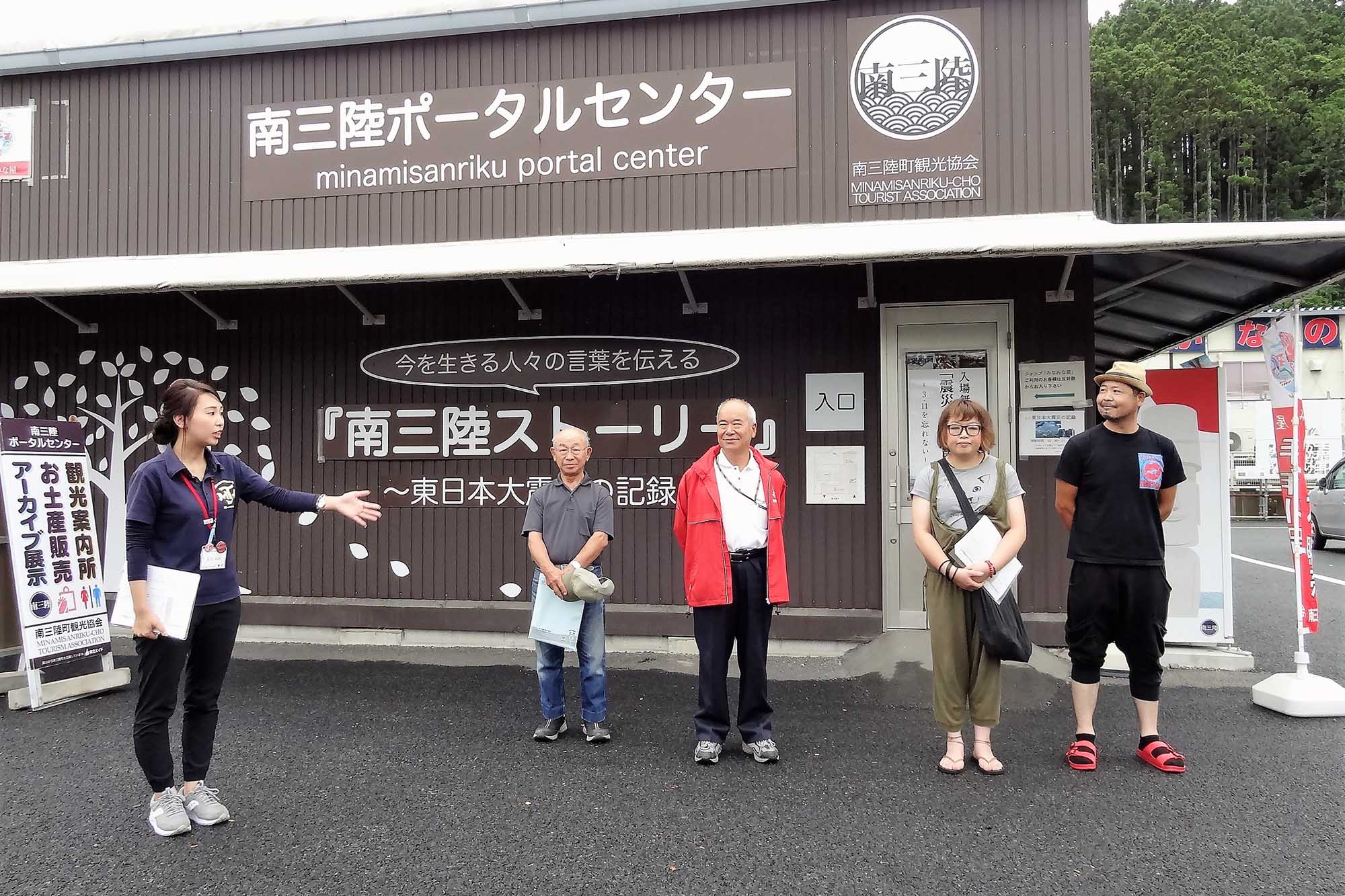 茶色の外壁に「南三陸ポータルセンター」と書かれた建物の前で、民泊家庭を紹介する女性が左手を広げて説明をしており、その横に首藤一雄さん、3人の方が並んで聞いている写真