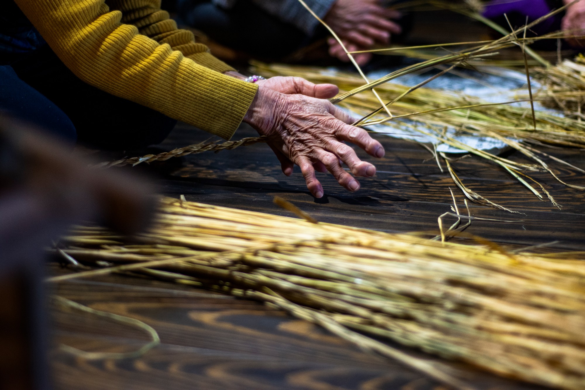 年配の人物が木の床に座り、手作業でわらをねじり縄を作っている手元を写した写真