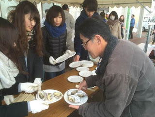 蓋を取ったカキが3つ乗った白い紙皿2つを軍手をした女性2人が男性客に渡しており、その隣では別の女性が紙皿をテーブルに並べている様子の写真