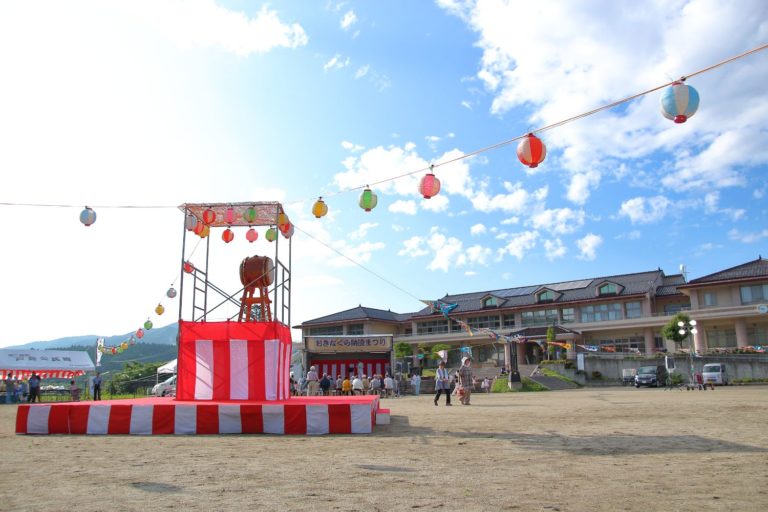 青空の下、紅白の縞模様のやぐらの上に太鼓が置かれ、周囲には色とりどりの提灯が飾られている、にぎやかな祭り会場の写真