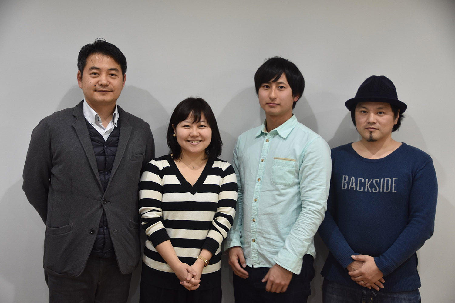 グレーの壁の前で、左からスーツ姿の男性、リポーターの女性、原田さん、ネイビーの帽子をかぶった男性が並んでいる写真