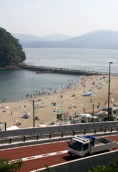 車が行き交う道路の先に広がる砂浜では、 色とりどりのパラソルやテントの下で、 多くの海水浴客で賑わっている様子の海水浴場の写真