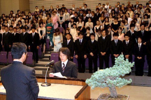 成人式の会場では、フォーマルな装いの新成人たちが客席で一斉に起立しており、壇上ではスーツを着た男性が代表として演台の前に立ち、誓いを述べている成人式の様子の写真