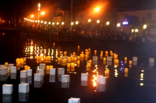 真っ暗な水面に灯籠が多数浮かび、その揺らめく光が水面に反射する幻想的な夜の情景と、奥の岸辺に並ぶ街灯と見物客のシルエットが写し出された流灯会の写真