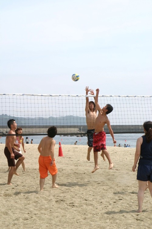 晴天の下、海水浴場に設置されたネット際で、水着姿の男女数人がビーチバレーのコート内に立ち、試合中に男性2人がネット際でボールを打ち合っている様子の写真