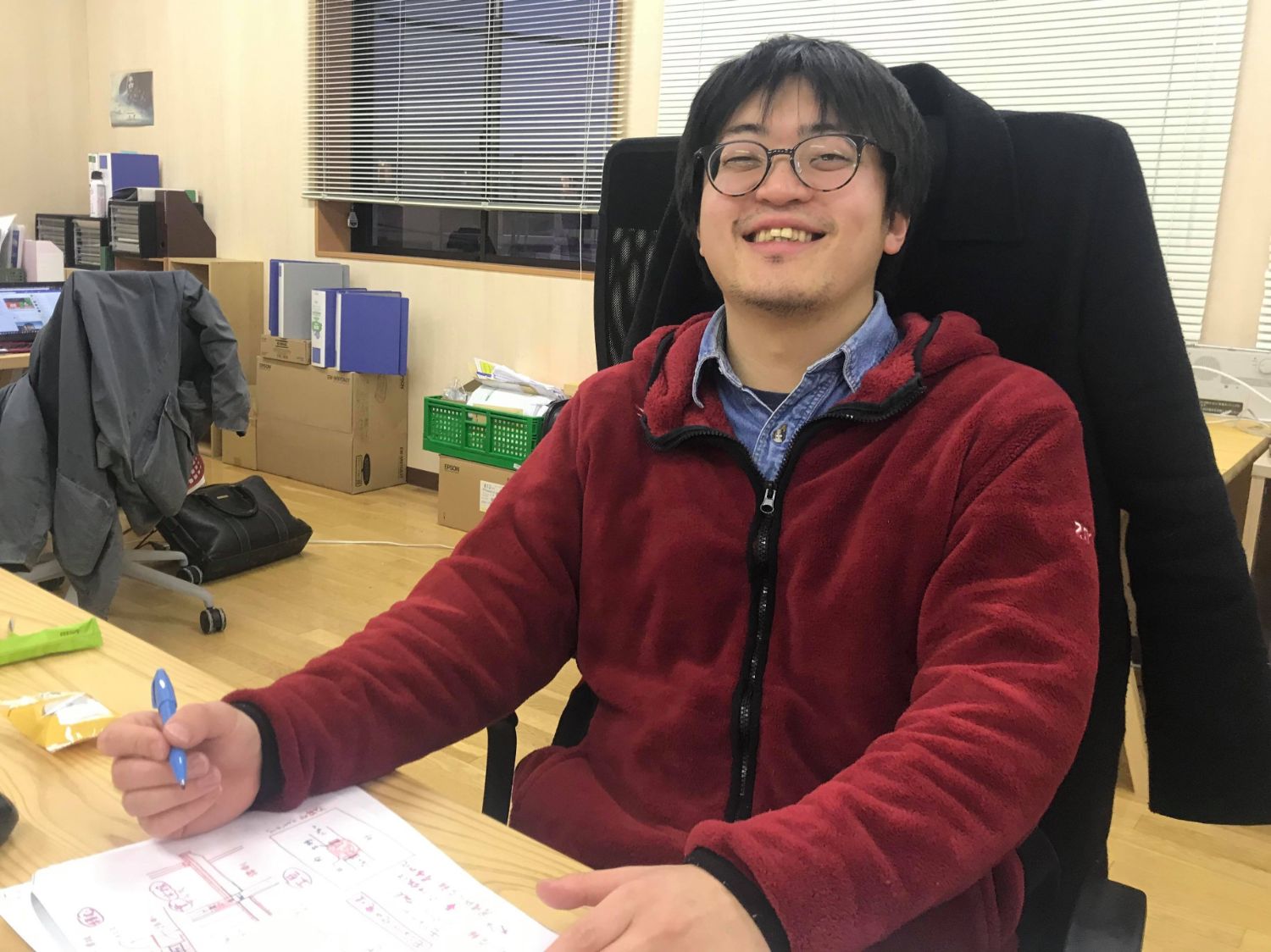 室内のオフィスで赤いフリースを着た男性が笑顔でペンを持ち、イラストの様なものが描かれた紙を置いた机の席に座っている写真