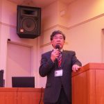 スピーカーが設置された室内会場の壇上で、スーツ姿の男性がマイクを持って話をしている写真