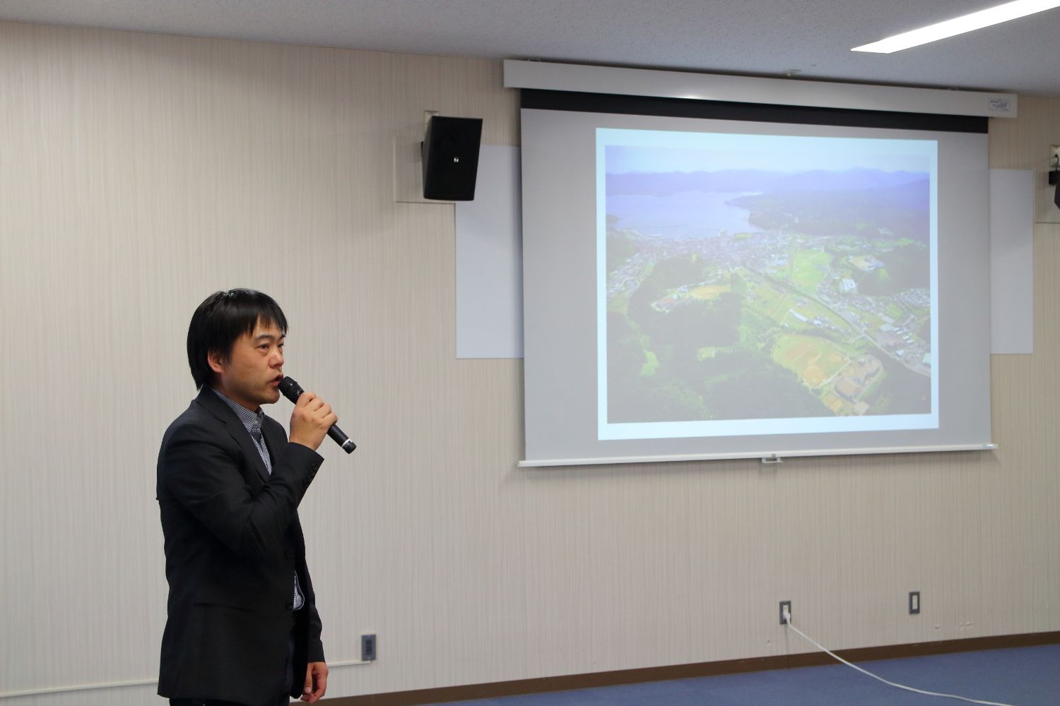 港町と海が映った航空写真を投影しているスクリーンの前に立つスーツを着た男性が、マイクを持って発表している様子の写真