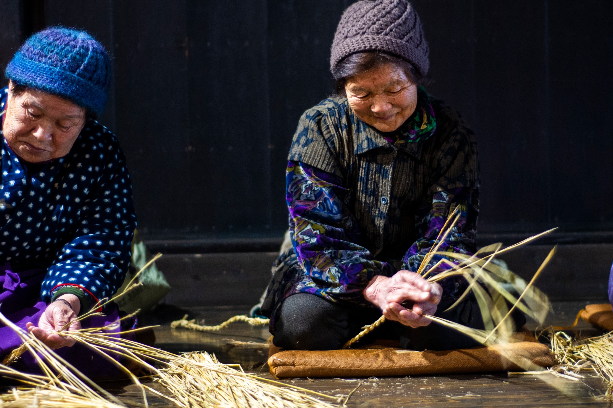 二人の人物が床に座りながら手作業で藁を編んでいる写真