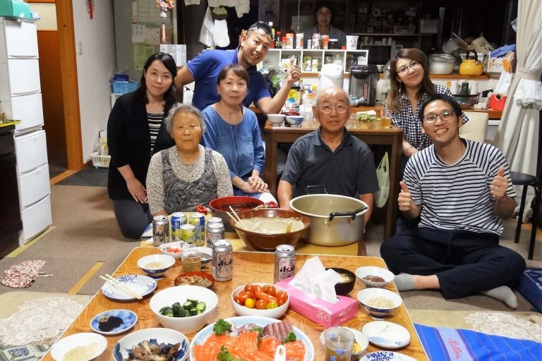 食卓に豪華な家庭料理を並んだ屋内の居間で、和子さんと一雄さん、他5人がくつろいで笑顔を見せている記念写真