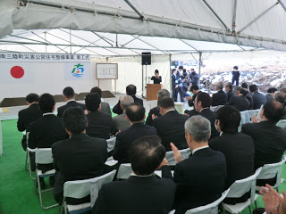 テント下に舞台が設置され、横断幕と国旗、町旗が掲げられ、多くの参加者が席についている災害公営住宅整備工事の着工式の様子の写真