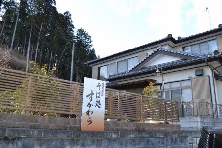 茶色の格子状の塀越しに「そば処すがわら」の看板が立っており、その背後に和風の民家が見える写真