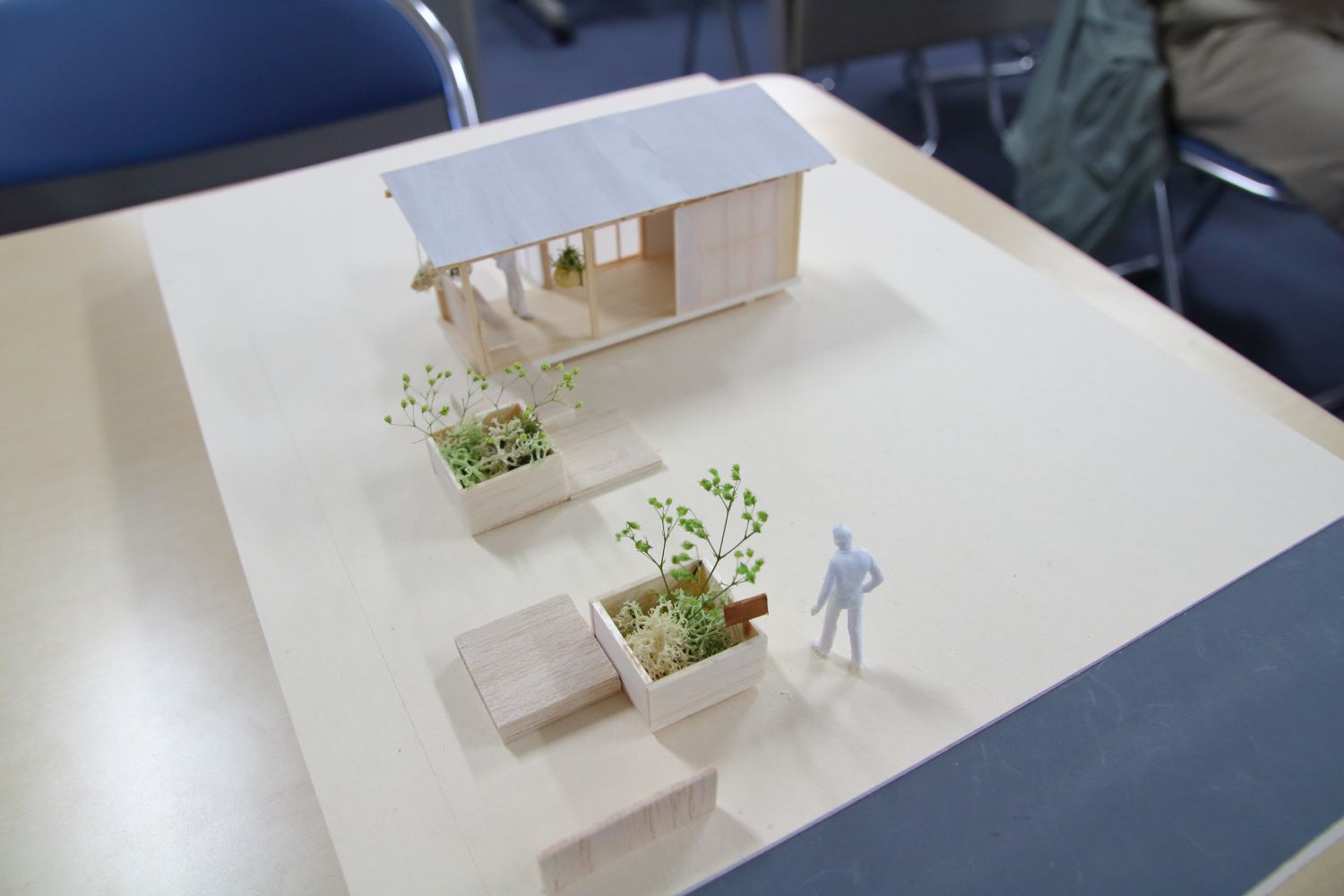 木で作られた小さな家と植木の模型がテーブルの上に置かれ、人の形をした白いフィギュアが傍に立っている建築模型の写真