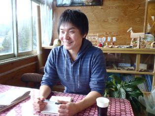 木製の棚に雑貨や植物が飾られた落ち着いた雰囲気の部屋で、テーブルに座っている大森さんが笑顔で写っている写真