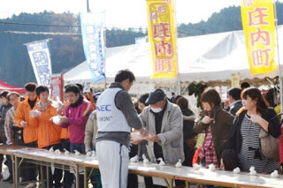 白いテントとのぼりが並び、たくさんの人でにぎわっている会場で、長く一列に並べられたテーブルの前に来場者が並び、NECと書かれた白いビブスをつけた男性が白いお椀を配っている写真