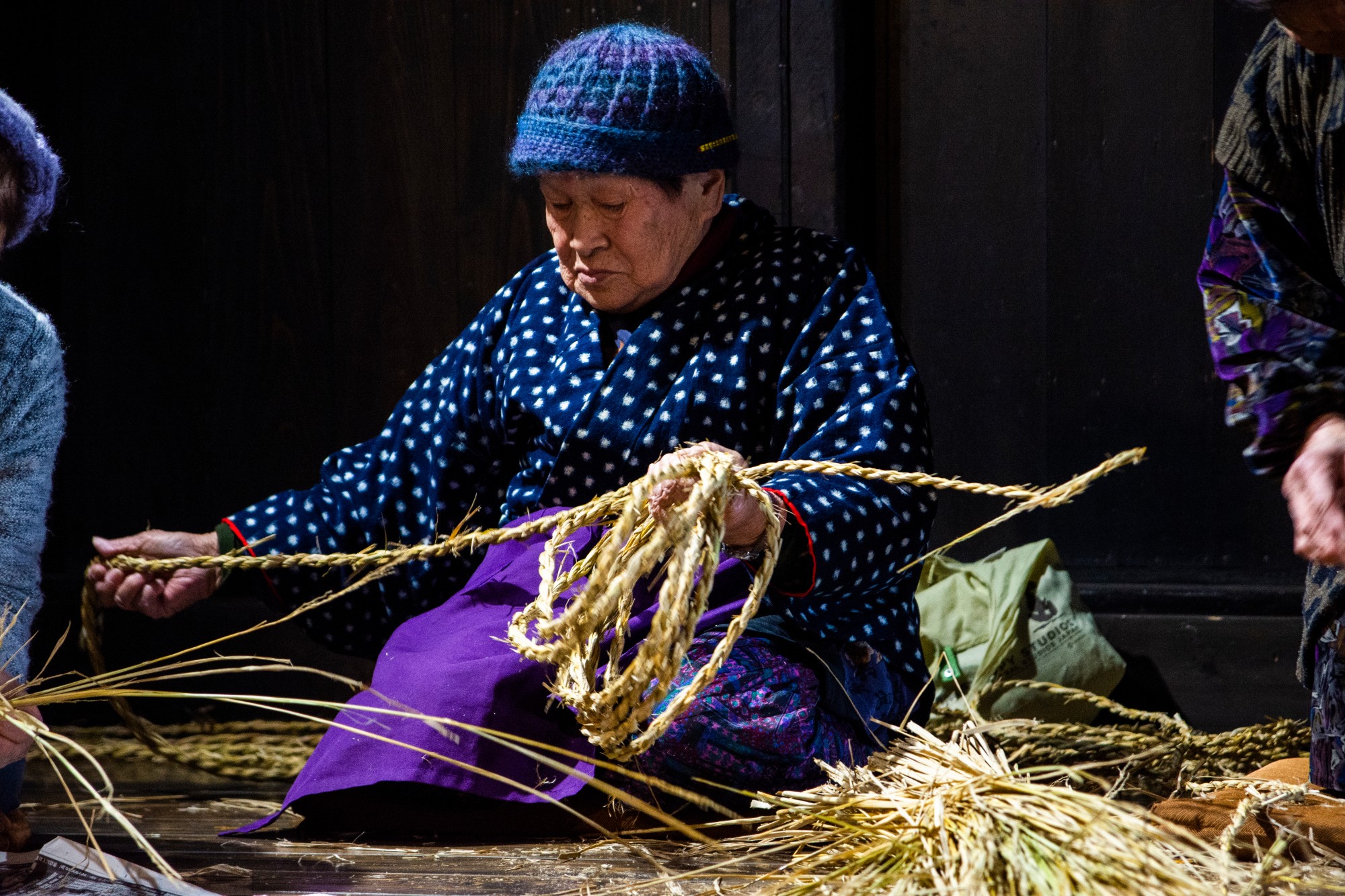 暗い背景の前で、青いニット帽を被った年配の女性が手作業で藁を編んでいる様子が写っている写真