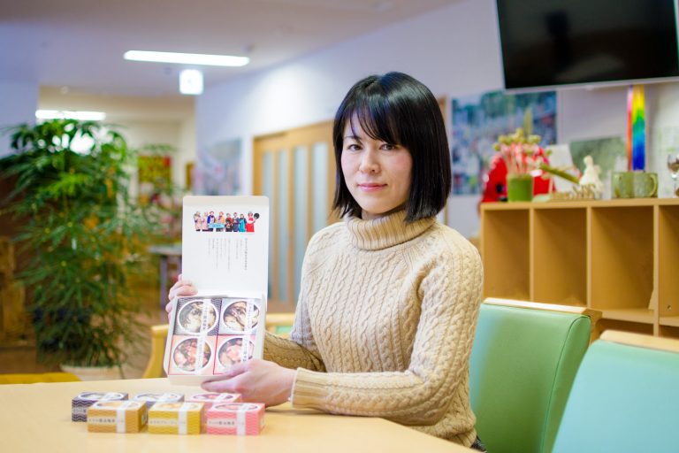 手造りのマグカップなどが飾られた棚の前のテーブルに、6種類の缶詰がテーブルに置かれ4種類の缶詰が入ったパッケージの箱を持って座る、白いセーターを着た中村さんの写真