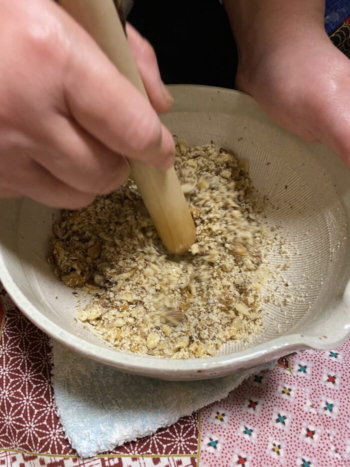 左手でしっかりとすりこぎを持ち、白いすり鉢の中でくるみを丁寧にすり潰している調理の様子の写真