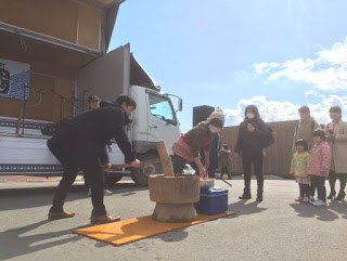 青空の下、荷台が開きマイクが並んで置かれた大型トラックの前で、木製の杵と臼を使って餅つきが行われ、親子連れなどが見物している写真