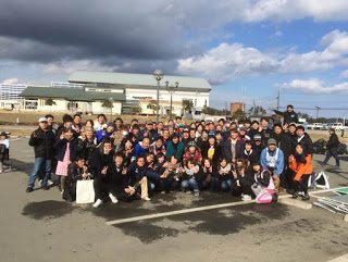 コンクリートの駐車場にたくさんの人々が集まり、何列にも並んで写った集合写真