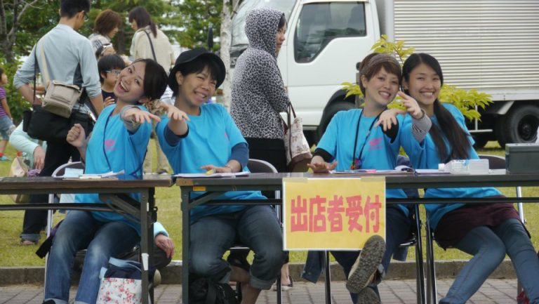 屋外イベントの出展者受付で青いTシャツを着た4人のスタッフが笑顔で座り、手を前に出してポーズをとっている写真