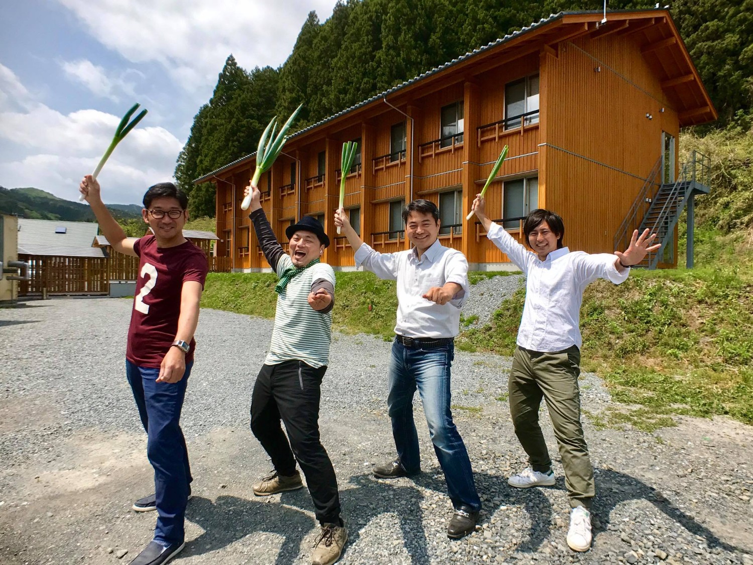 木々に囲まれた2階建ての木造の建物を背景に、屋外で4人の男性が長ネギを手に持ってポーズを決めながら笑顔で並んでいる写真