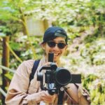 黒いキャップを被り、サングラスをかけた男性が笑顔でカメラを構えている写真