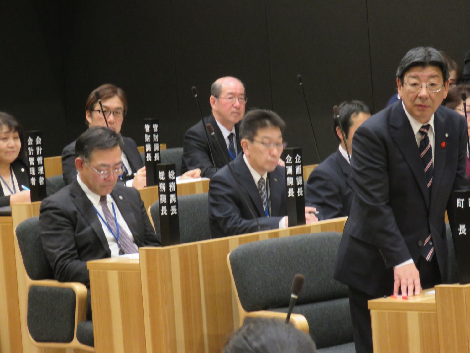 「町長」と書かれた議席札がある席で、佐藤町長が起立し話をしている様子の写真
