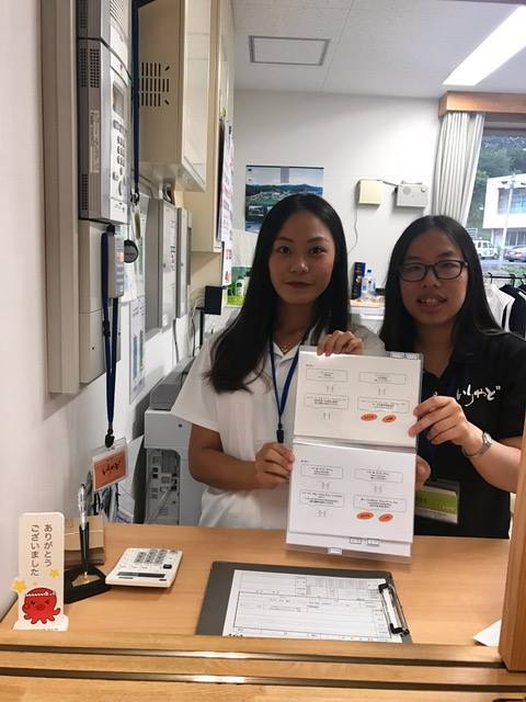 受付カウンターの奥で白いシャツの女性と黒い服の女性が、ファイルに入った資料を手に持って並んでいる写真