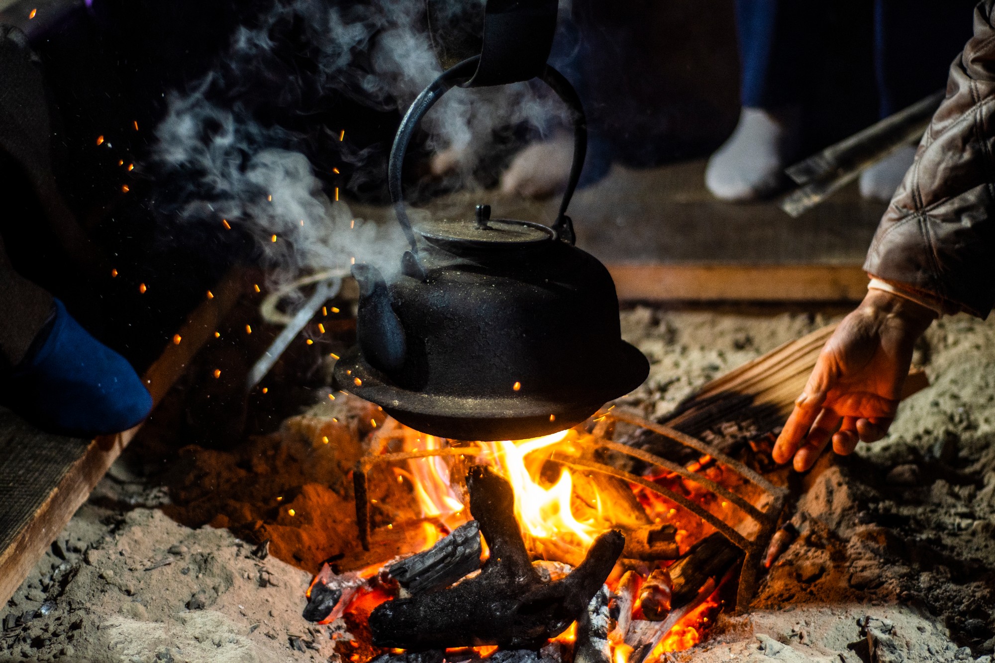 囲炉裏の火にかけられた鉄瓶から湯気が立ち上る様子と、右手に火箸を持って炭を調整している人の手元が写っている写真