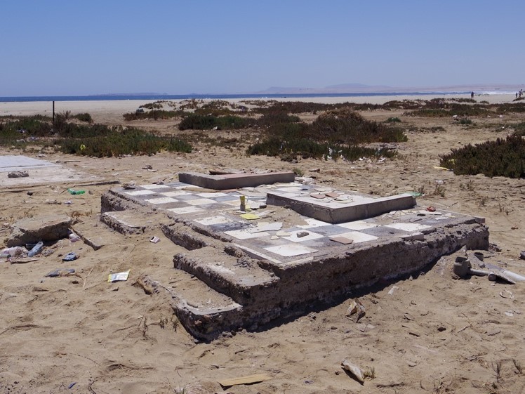 砂浜にあるタイル張りの基礎部分が残された廃墟が写っており、周囲には低い草木が点在し、遠くには青い海が広がっている写真