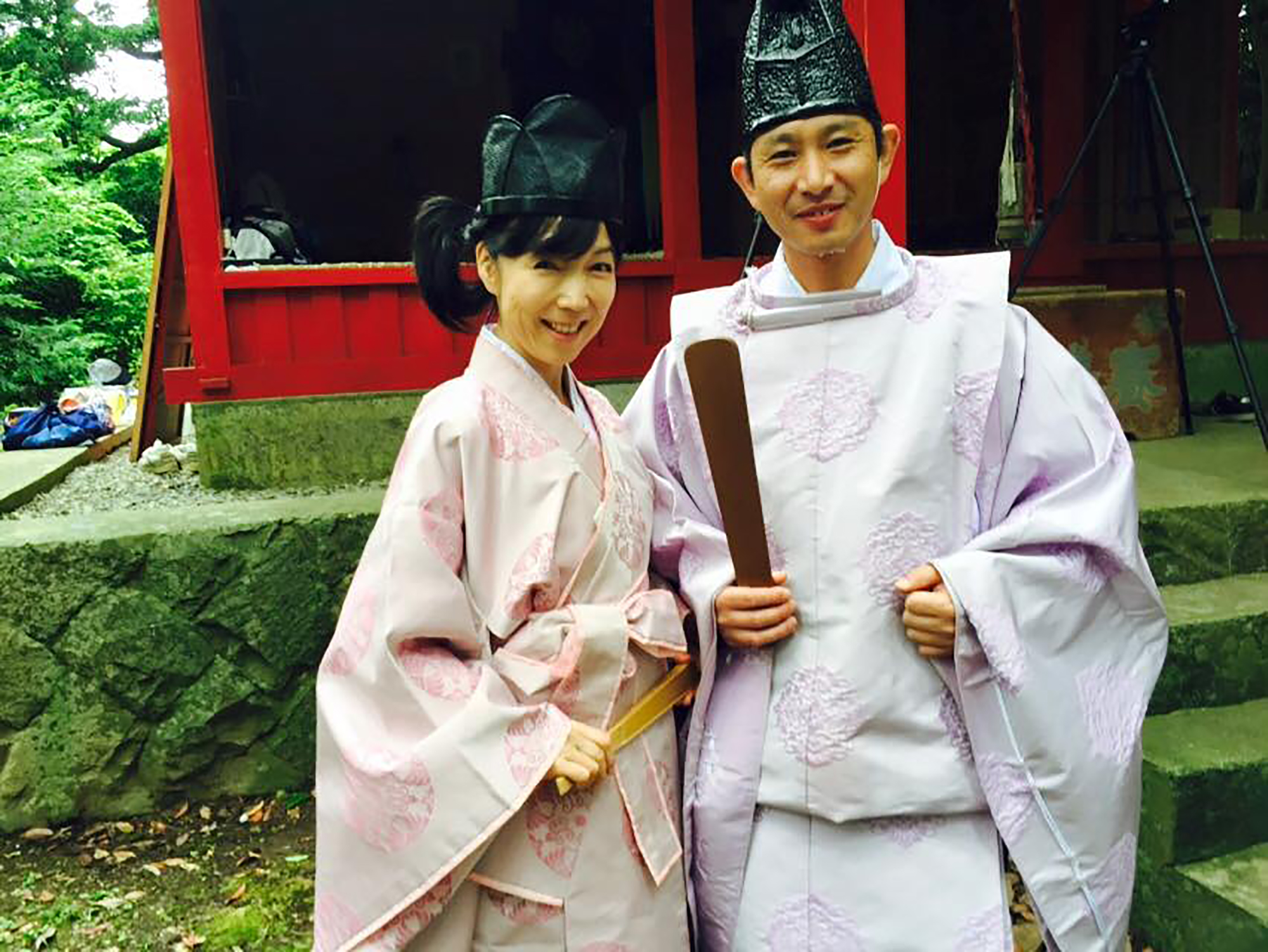 伝統的な装束を着て黒い冠帽子をかぶった奥さまの工藤真弓さんと工藤庄悦さんが、屋外の石段の前で並んで笑顔を見せている写真