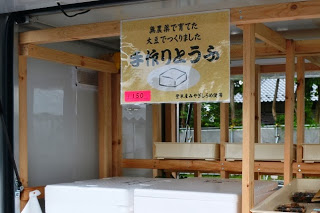 「手作りとうふ」と書かれた黄色い看板の下に保冷容器が2つ並べて置かれている豆腐販売スペースの写真