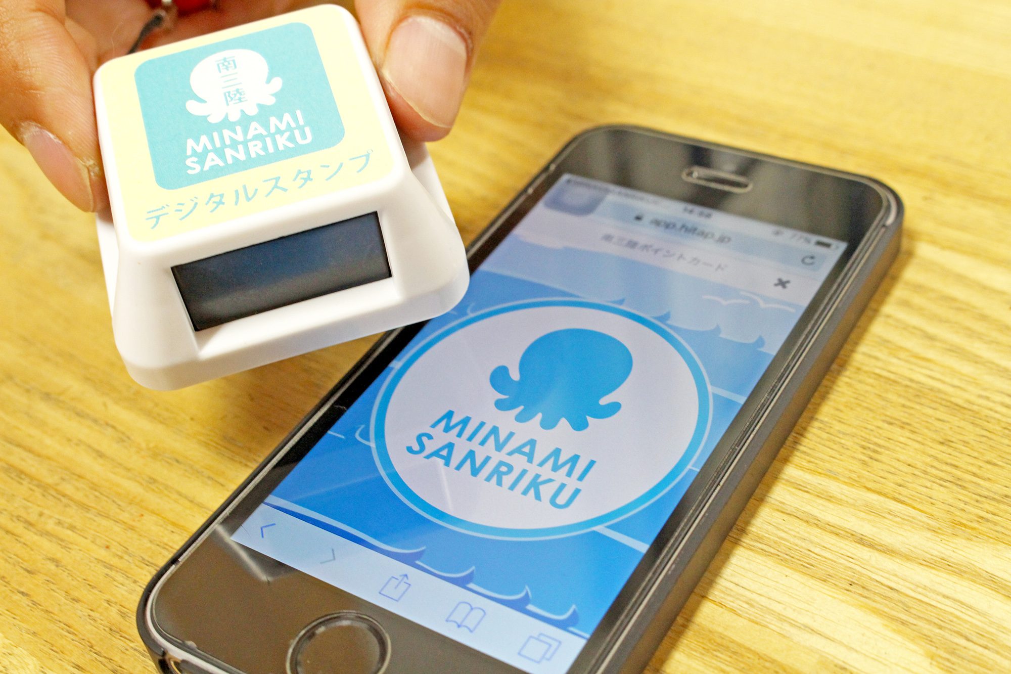 スマートフォンの画面に表示されたタコのマークと「MINAMI SANRIKU」のロゴマークに、白くて四角い電子スタンプをかざしている手元が写っており、スタンプにはタコのマークと「南三陸 MINAMI SANRIKU デジタルスタンプ」と書かれている様子が映っている写真