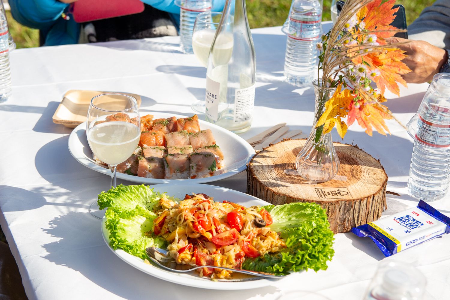 白いテーブルクロスが敷かれた屋外のテーブルの上に、「秋鮭の洋風巻きずし」「ホヤのサラダ」の料理と飲み物が並んでいる写真