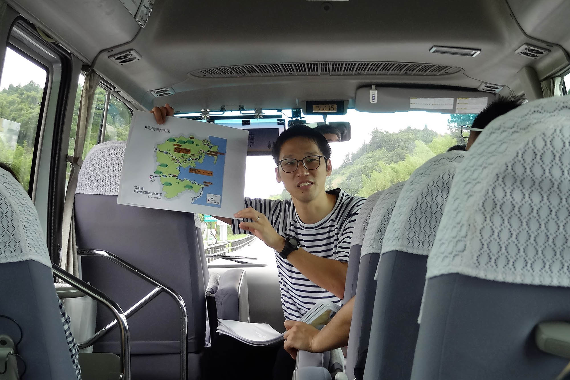 マイクロバスの車内で、眼鏡をかけた男性が地図を広げて説明している写真