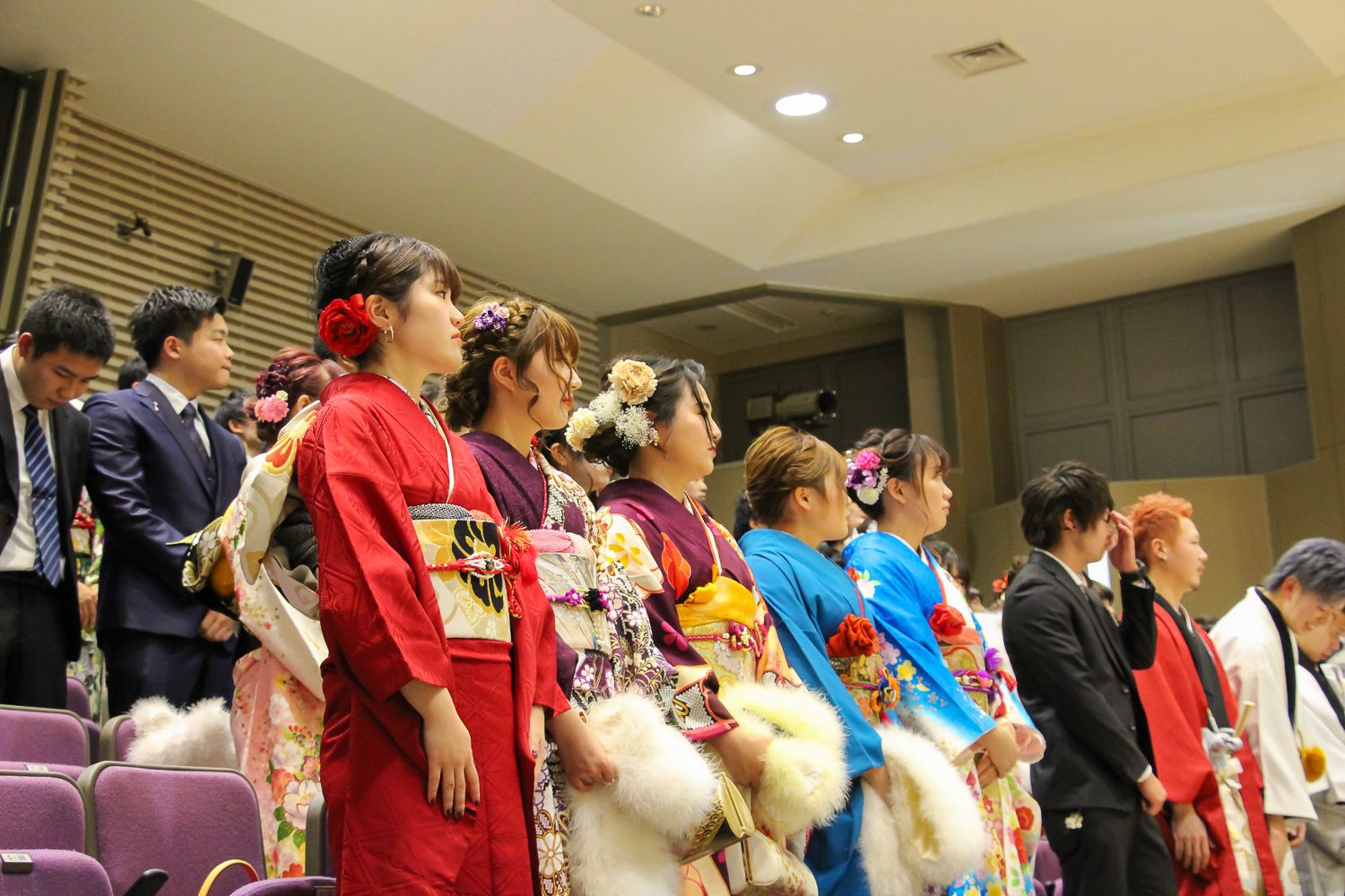 色とりどりの振袖や落ち着いた色のスーツを身につけた新成人たちが代表挨拶を聞いている様子の写真