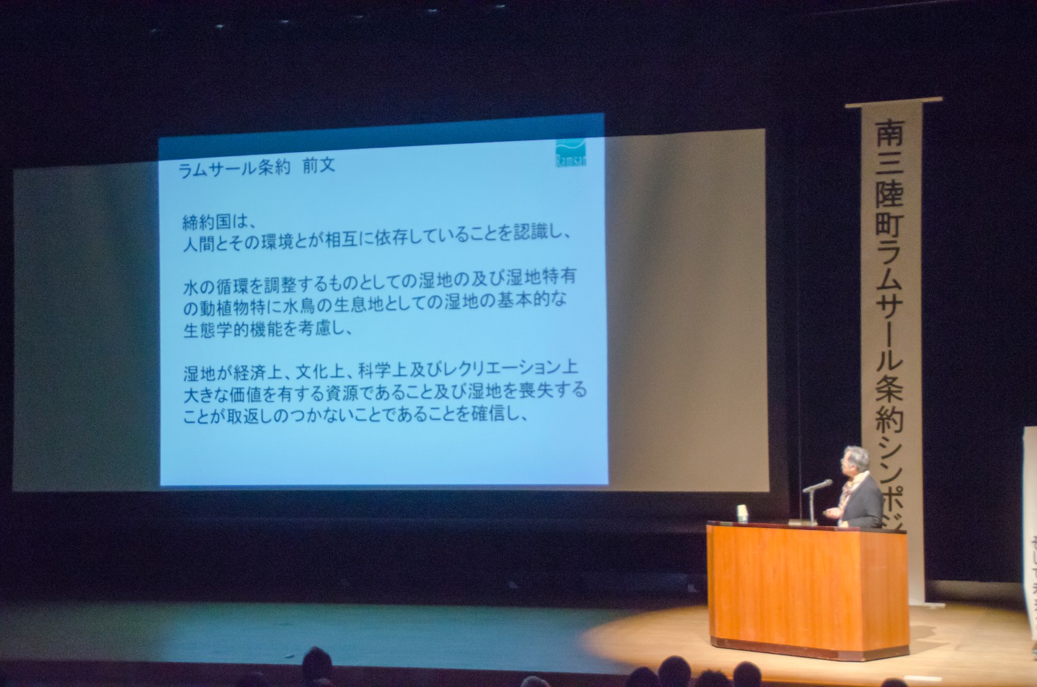 舞台のスクリーンにラムサール条約の全文を投影しながら講演する人物を写した講演の様子の写真