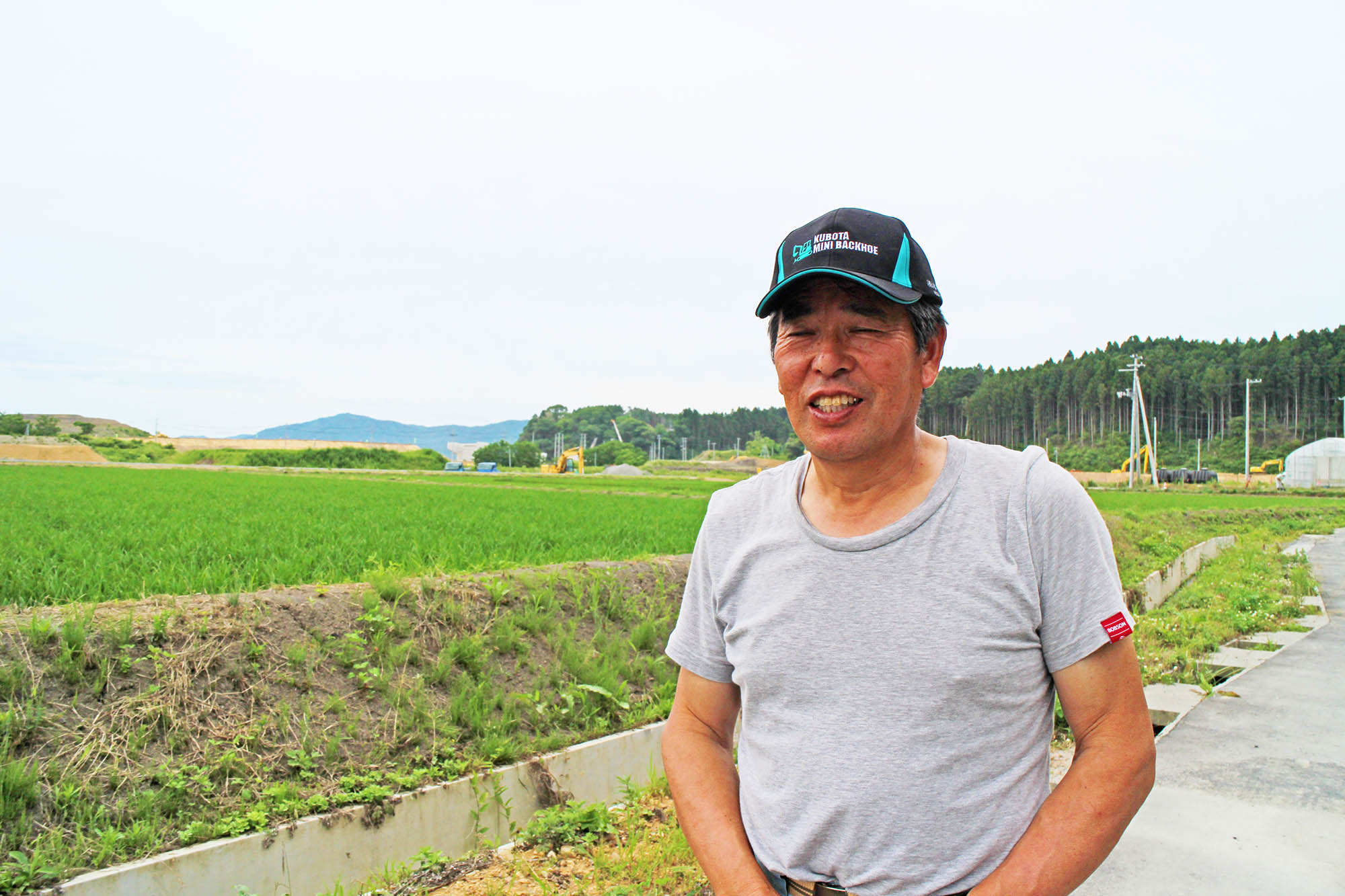 畑のそばの舗装された道に立ち、笑顔で写っている西城善昭さんの写真