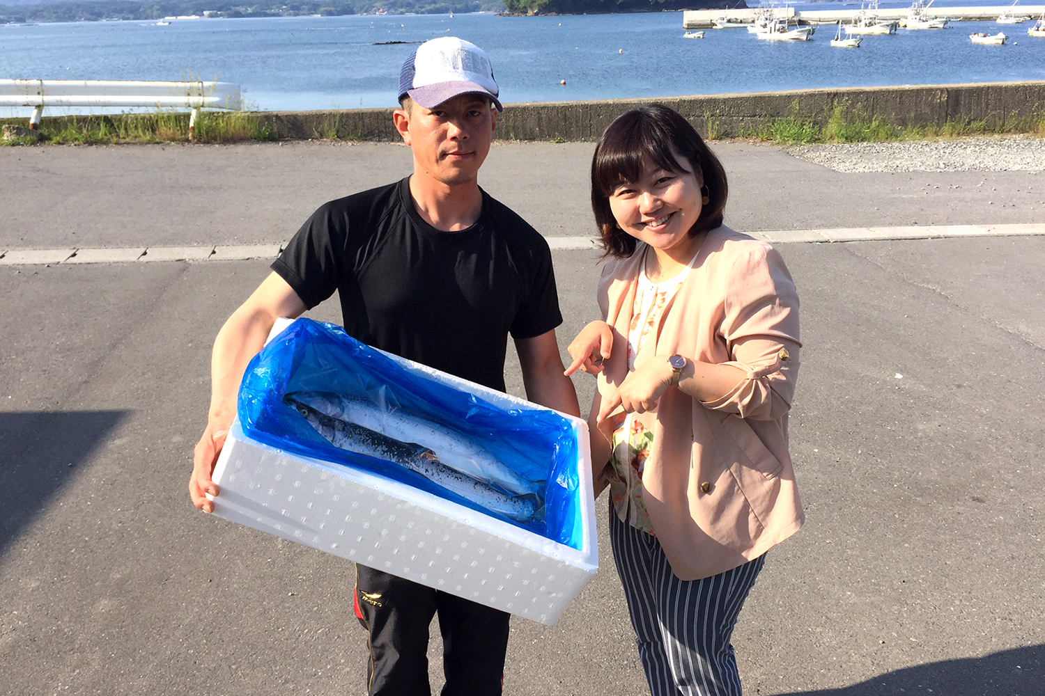 海沿いの道路を背景に、銀鮭が入った白い発泡スチロールの箱を抱えた須藤さんと女性リポーターが並んで立っている写真