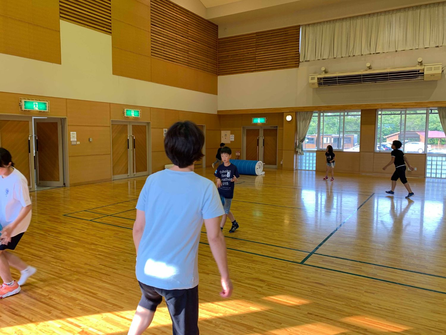 広々とした木の床の体育館で、複数の子どもたちが遊んでいる様子をTシャツ姿の女性が見ている後ろ姿の写真
