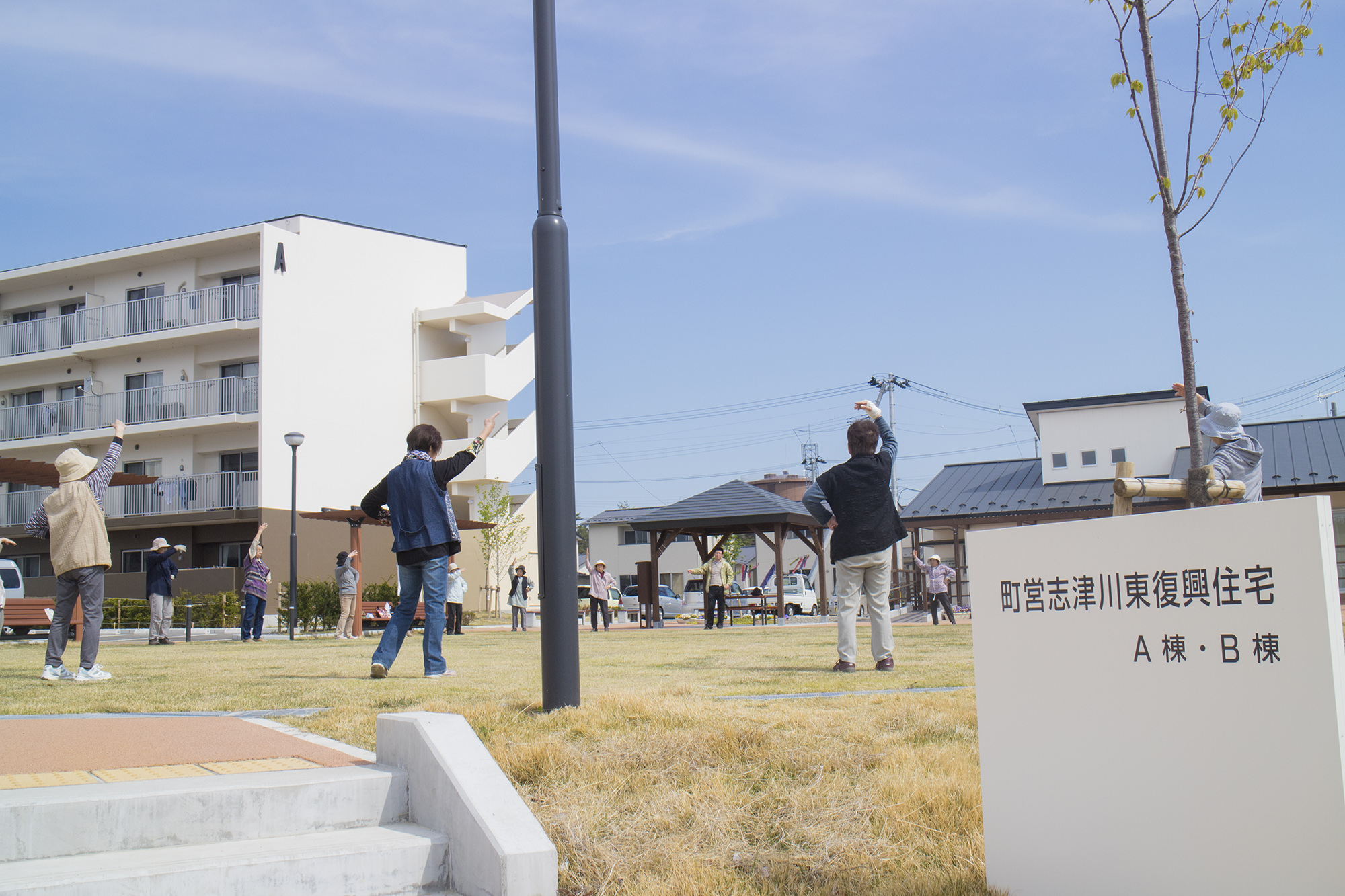 青空の下「町営志津川東復興住宅 A棟・B棟」の前の広場で、地域の方々が屋外で体操を行っている様子を写した写真