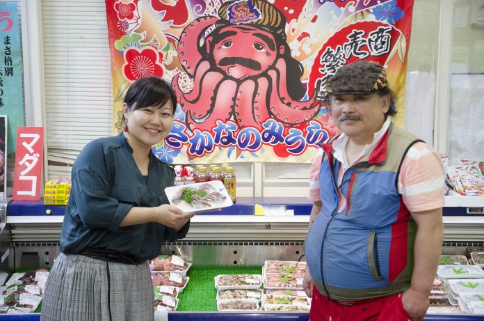 タコのイラストが描かれた大漁旗が掲げられたオープンタイプの冷蔵ショーケースの前で、リポーターの女性がパックに入った魚の刺身を笑顔でカメラに見せ、横で帽子をかぶったベスト姿の三浦保志社長が並んで立っている写真