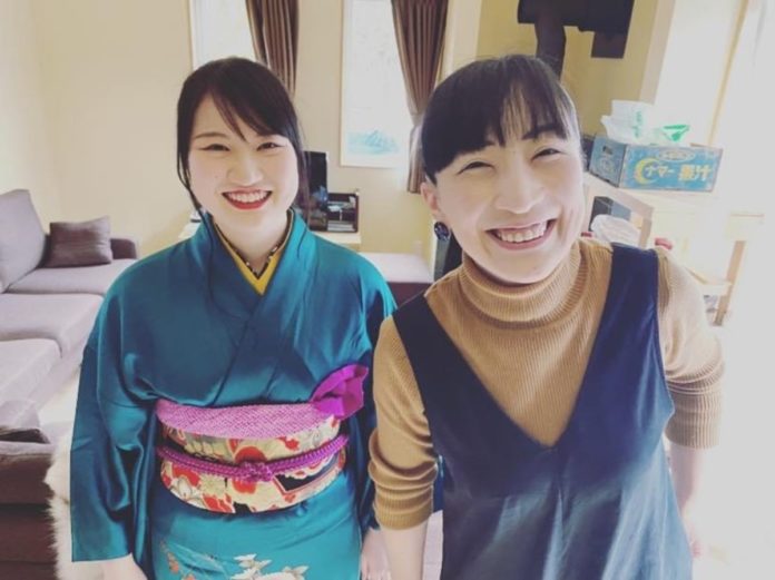 緑色の着物を着た女性とセーターとジャンパースカートを着た女性が室内で笑顔を見せながら並んで立っている写真