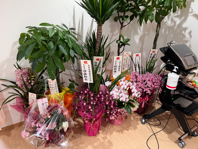 院内の一角に観葉植物や胡蝶蘭など多数の祝い花が並べられており、花に添えられた札には関係者の名前と祝辞が書かれている写真