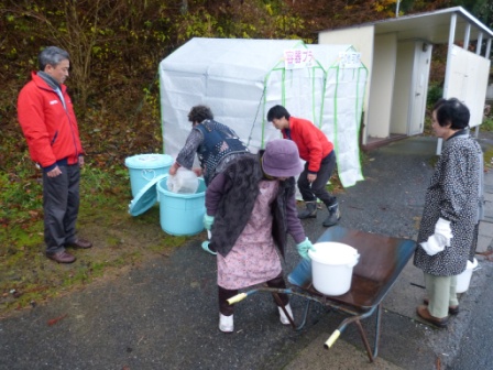 屋外の分別ステーション付近で、複数の人々がバケツを持ってごみを運び、分別作業を行っている、ごみ分別活動の写真