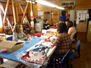 木造の作業場の中で、Yes工房で働く4人の女性がテーブルを囲み、布製品や箱を使って手作業をしている様子の写真